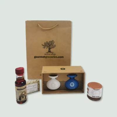 A brown paper gift bag with the logo and text "gourmet.wearedoubledot.com" sits behind a selection of gourmet products. These include a small bottle of Commandaria red wine with a red cap, a bar of soap in green and white packaging, a box containing a white ceramic bottle of extra virgin olive oil and a blue ceramic bottle of balsamic vinegar, and a jar of pine honey.