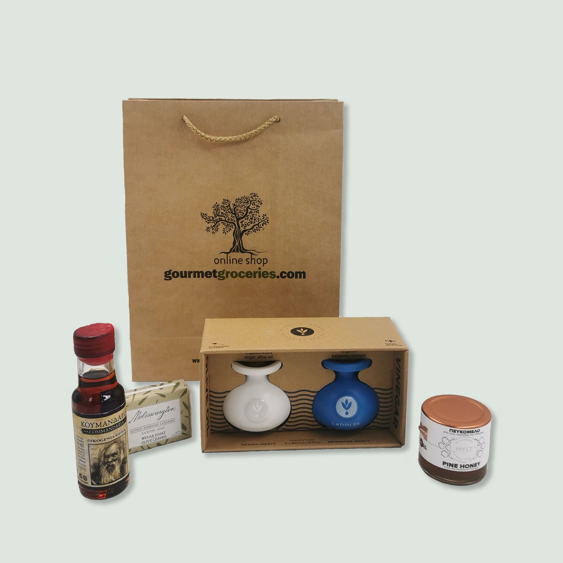 A brown paper gift bag with the logo and text "gourmet.wearedoubledot.com" sits behind a selection of gourmet products. These include a small bottle of Commandaria red wine with a red cap, a bar of soap in green and white packaging, a box containing a white ceramic bottle of extra virgin olive oil and a blue ceramic bottle of balsamic vinegar, and a jar of pine honey.