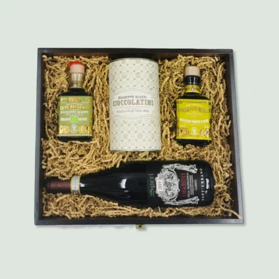 A wooden gift box containing gourmet items arranged neatly in wood wool. The items include a bottle of balsamic vinegar, a tin of chocolate, a bottle of olive oil, and a bottle of red wine.