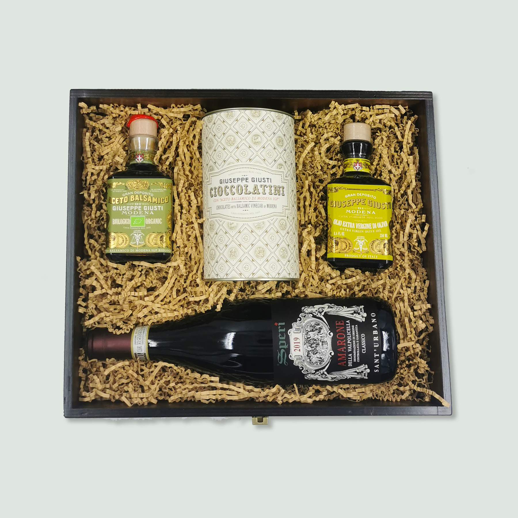 A wooden gift box containing gourmet items arranged neatly in wood wool. The items include a bottle of balsamic vinegar, a tin of chocolate, a bottle of olive oil, and a bottle of red wine.