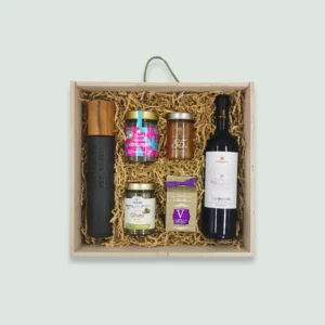 A wooden gift box containing various gourmet items arranged neatly in wood wool. The items include a bottle of olive oil, a jar of hyssop herbs, a jar of mandarin marmalade, a jar of olives, a package of Greek honey with mango and walnuts, and a bottle of red wine.