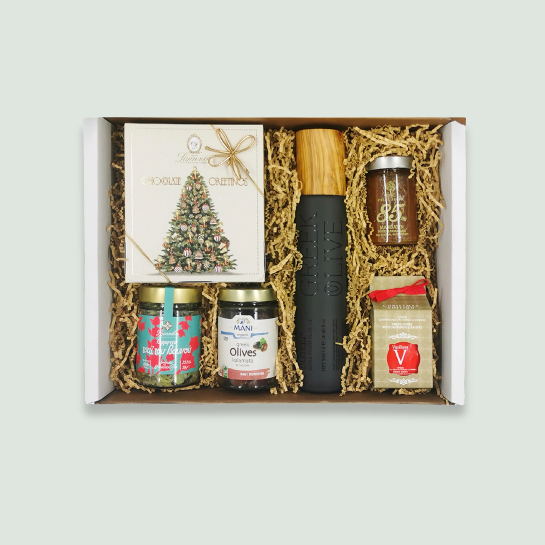 A gift box containing a bottle of Greek olive oil, a Christmas-themed box of chocolates, a jar of mountain tea, a jar of Kalamata olives, a jar of mandarin marmalade, and a jar of Greek honey with cinnamon and raisins. The items are nestled in wood wool within a cardboard box.