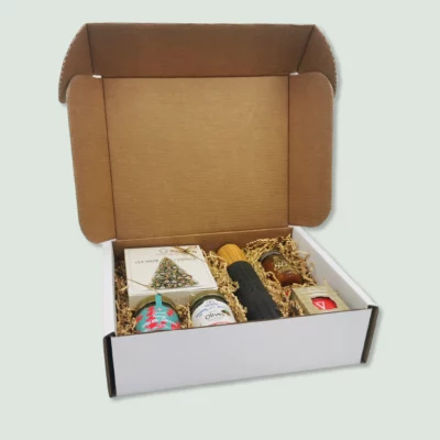A front angle of a gift box containing a bottle of Greek olive oil, a Christmas-themed box of chocolates, a jar of mountain tea, a jar of Kalamata olives, a jar of mandarin marmalade, and a jar of Greek honey with cinnamon and raisins. The items are nestled in wood wool within a cardboard box.