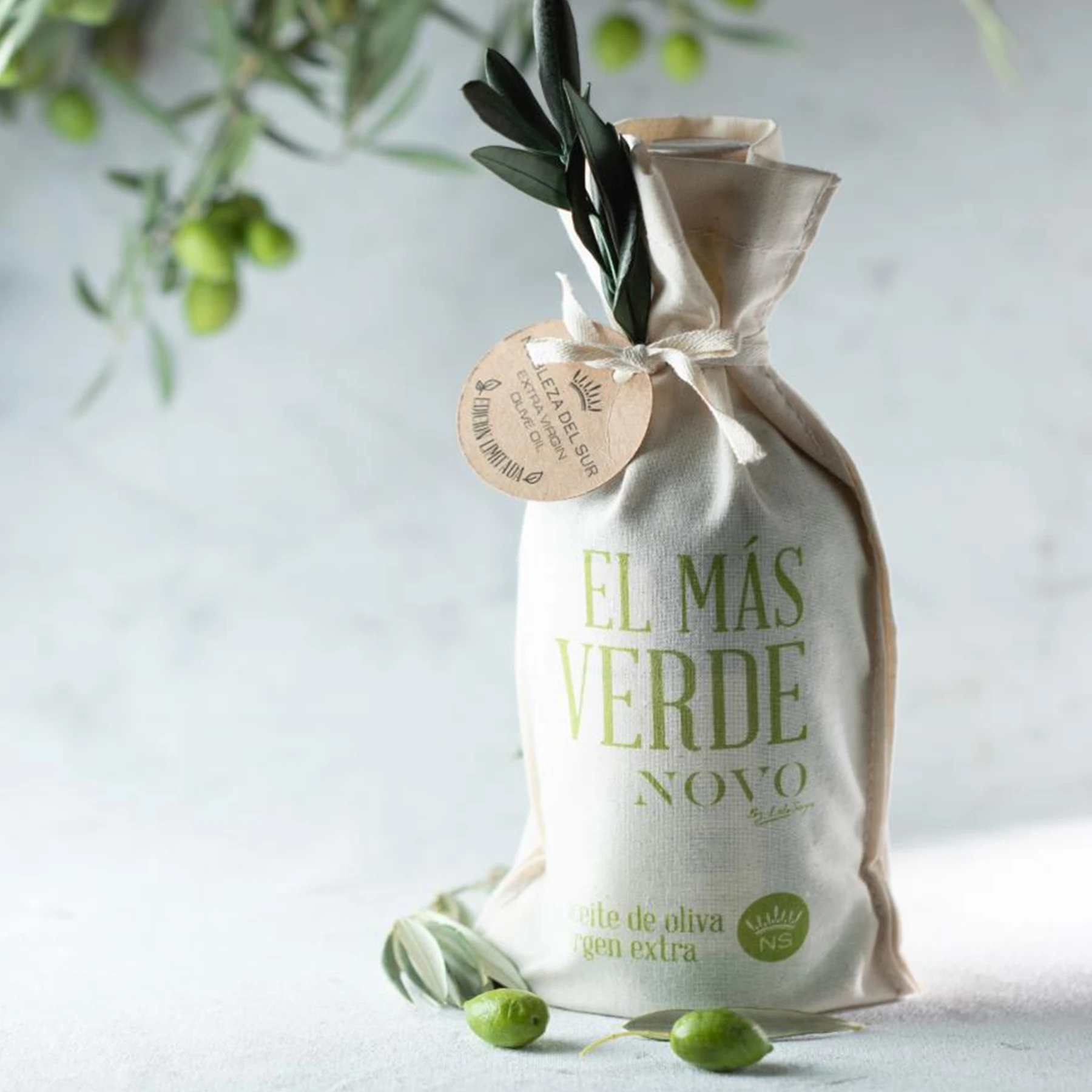 A bottle of El Más Verde Novo extra virgin olive oil positioned next to a bowl of fresh olives, emphasizing the oil's quality and origin.