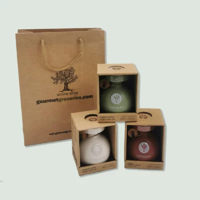Three boxed ceramic bottles of Ladolea organic products displayed in front of a brown paper gift bag with a rope handle. The bag is branded with the logo "gourmet.wearedoubledot.com." The bottles include organic extra virgin olive oil in white and green bottles, and organic balsamic vinegar with bergamot in a red bottle.