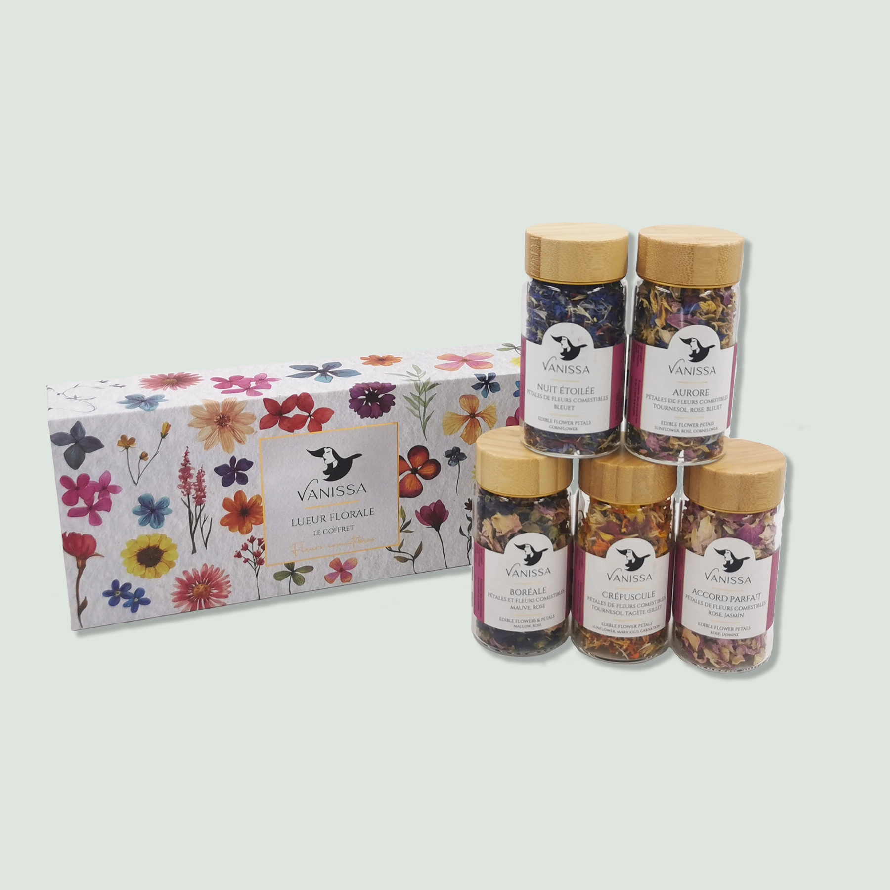 A box with five jars of mixed herbs by Vanissa, labeled "Lueur Florale." The jars have golden lids and contain colorful blends of edible flowers and herbs. The box features a floral design with various flowers illustrated on it, and the jars are arranged outside the box for display.