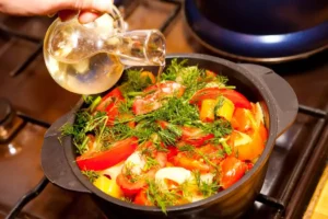 An image of a person pouring olive oil from a clear glass bottle into a pan of mixed vegetables on a stovetop.