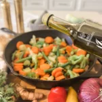 Pouring olive oil into a pan filled with colorful vegetables and herbs.